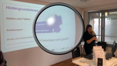 Ramona Armbrust steht vor einer Wand auf der eine Bildschirmpräsentation ist. Auf der Folie steht "Hintergrundwissen, Zahlen und Fakten". In der Mitte des Bildes ist eine große Lupe eingefügt, die einen Teil des Fotos vergrößert.