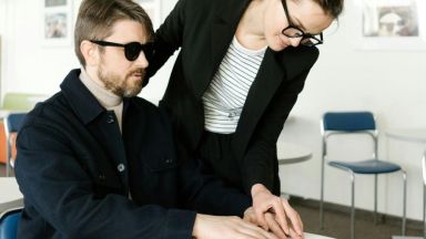 In einem Büro: Eine Frau zeigt einem offenbar blinden Mann etwas in einem aufgeschlagenen Buch. Dazu führt sie seine Hand. 